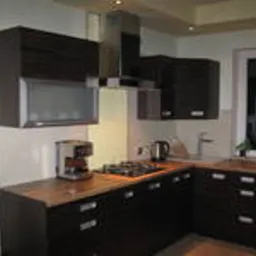 Ciemna, narożna zabudowa kuchenna z drewnianym blatem, widoczny ekspres do kawy i płyta gazowa, oświetlenie punktowe podszafkowe, okno w tle.